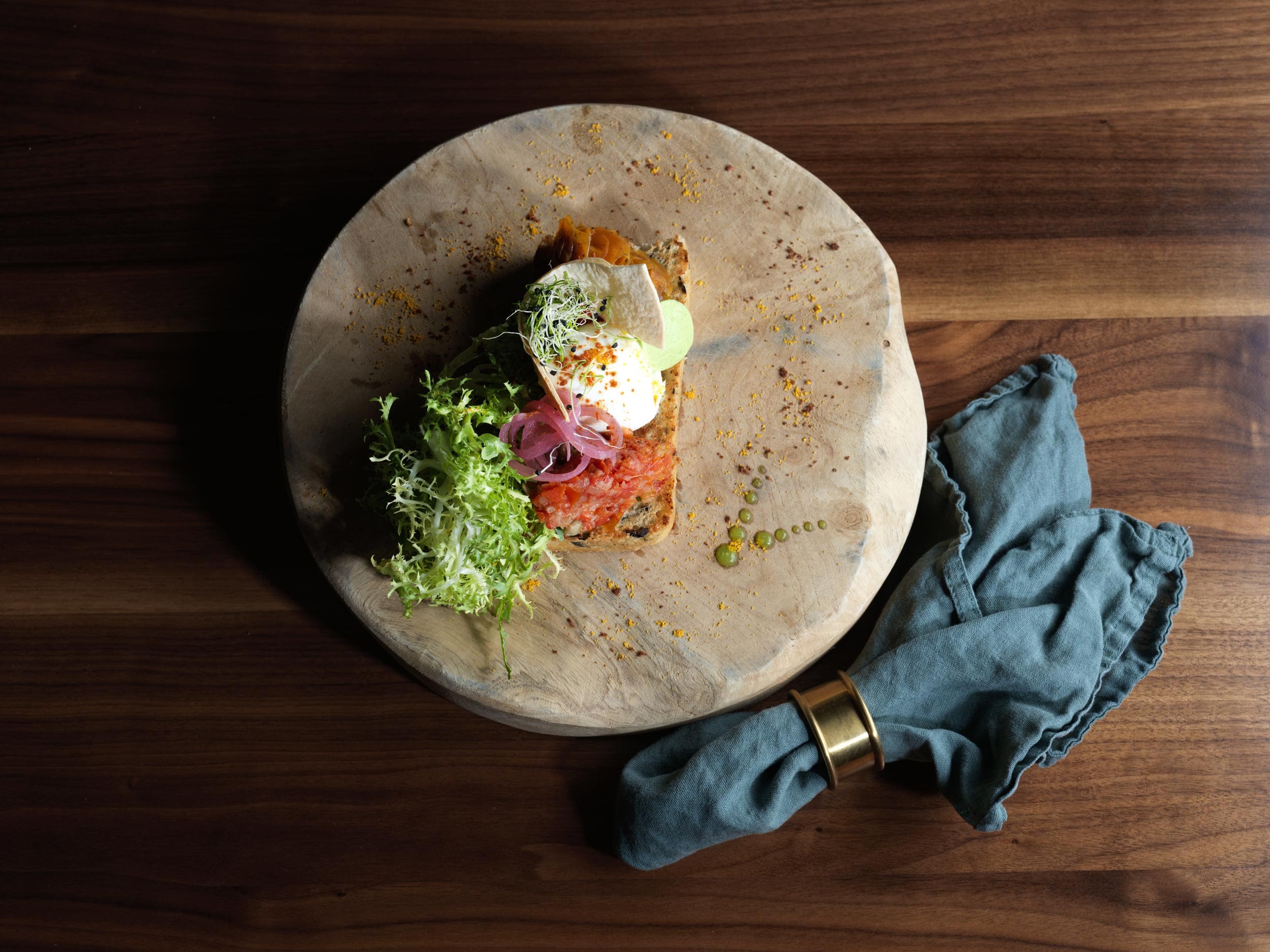 Auf einem Holzteller liegt Toast im Restaurant-Stil mit pochiertem Ei, Tomaten, Gemüse, eingelegten Zwiebeln und Microgreens. Friséesalat steht daneben. Daneben liegt eine blaue Stoffserviette mit goldenem Ring – perfekt zum Wein.