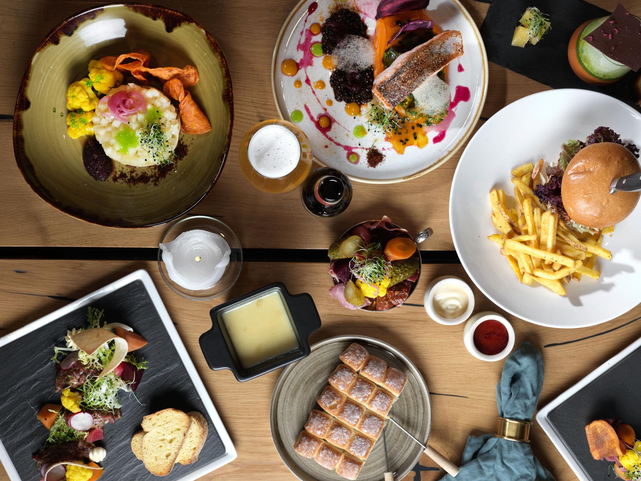 Eine Draufsicht auf einen Restauranttisch, der mit verschiedenen Gourmetgerichten gedeckt ist, darunter ein Burger mit Pommes, Lachs, Dessert, Soßen, Brot, Gemüse und Craft Beer – perfekt zum Wein.
