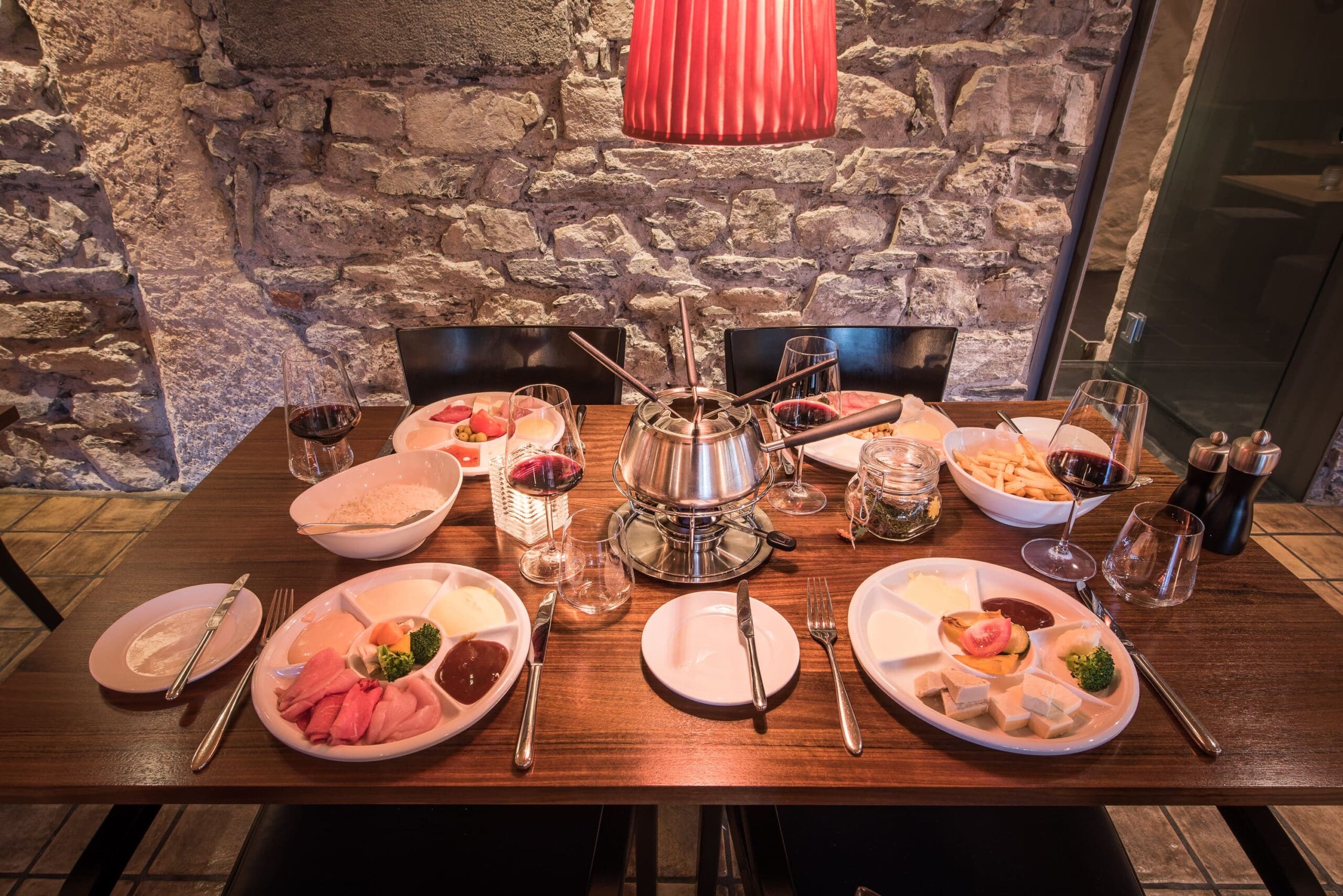 Ein für vier Personen gedeckter Restauranttisch mit Tellern mit Aufschnitt, Soßen, Käse, Brot und einem Fonduetopf in der Mitte, umgeben von Rotweingläsern, unter einer roten Hängelampe an einer Steinwand.
