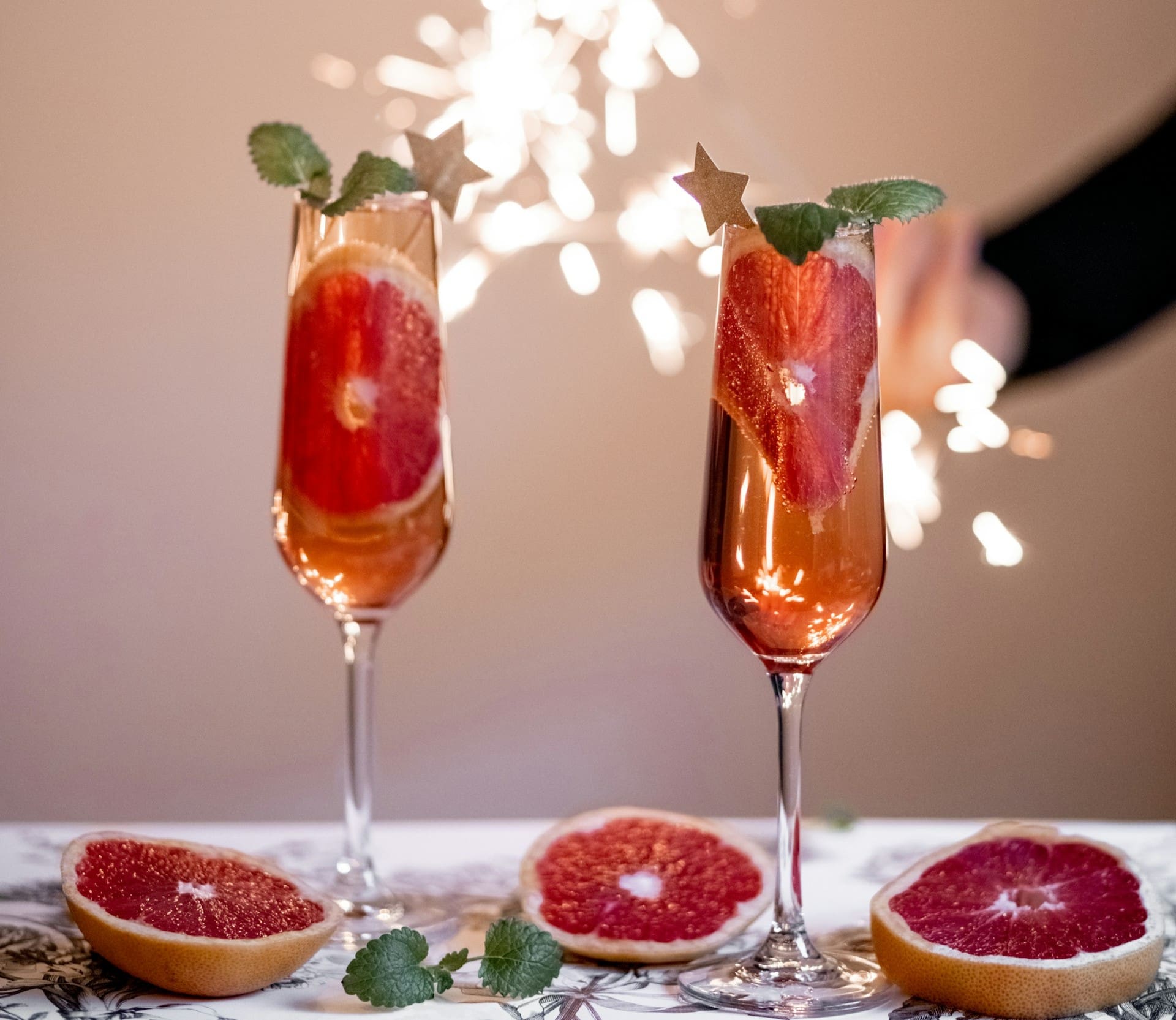 Deux coupes de champagne avec des tranches de pamplemousse et des feuilles de menthe, garnies de pics étoilés dorés, sont posées sur une table avec des moitiés de pamplemousse. À l'arrière-plan, une main tient un cierge magique allumé, créant ainsi une atmosphère festive.