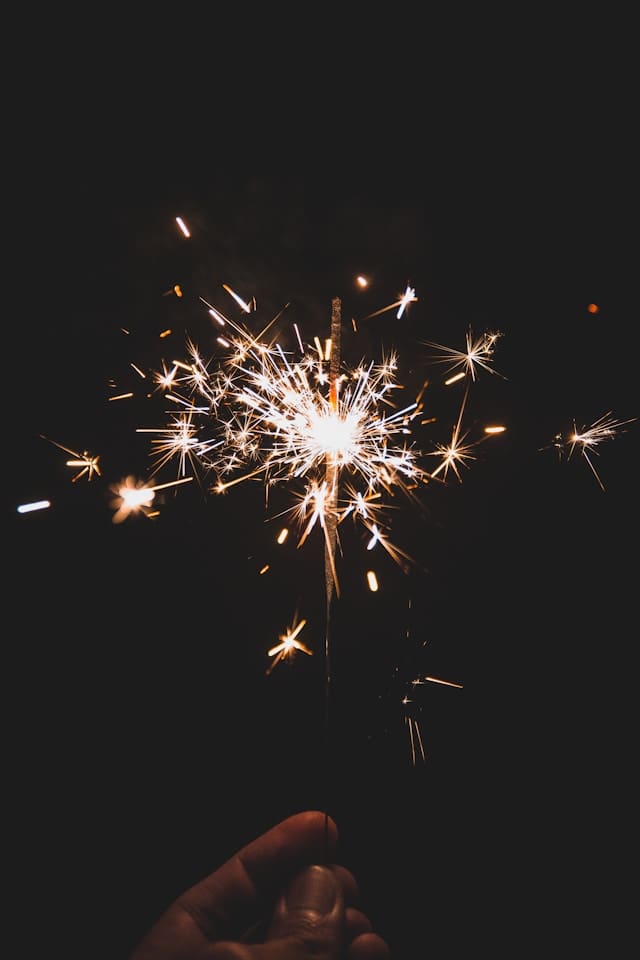 A hand holds a lit sparkler in the dark, with bright sparks radiating outward against a black background.