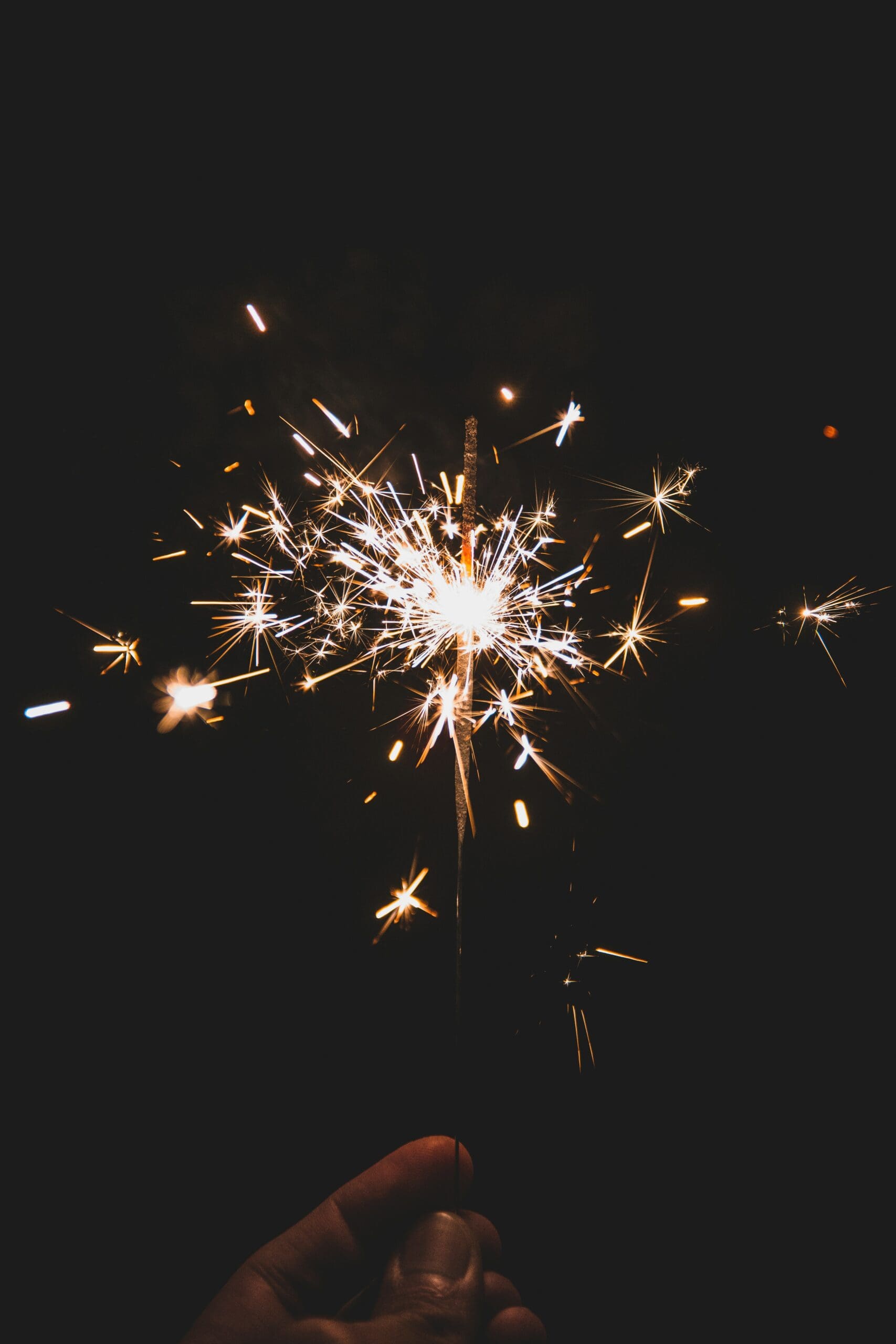 A hand holding a lit sparkler, with bright, glowing sparks radiating in the dark, black background.